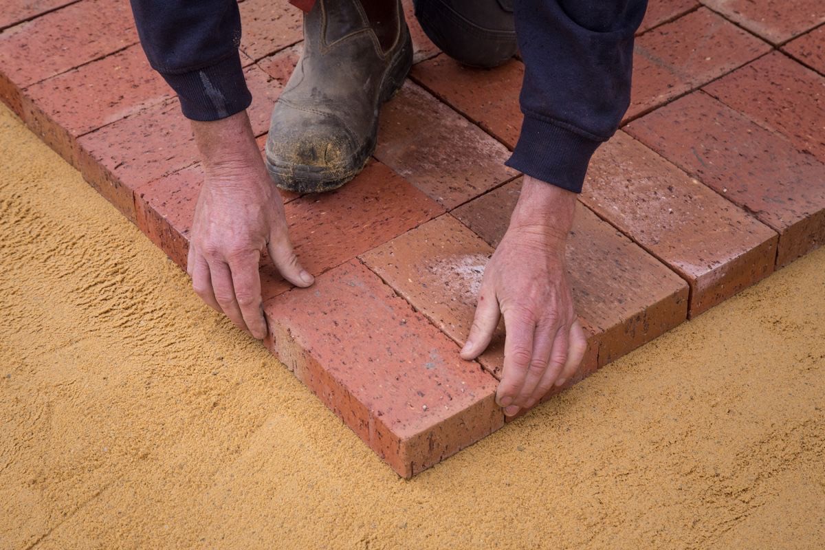 Hands setting clay brick pavers tight against each other on a screeded sand bedding layer